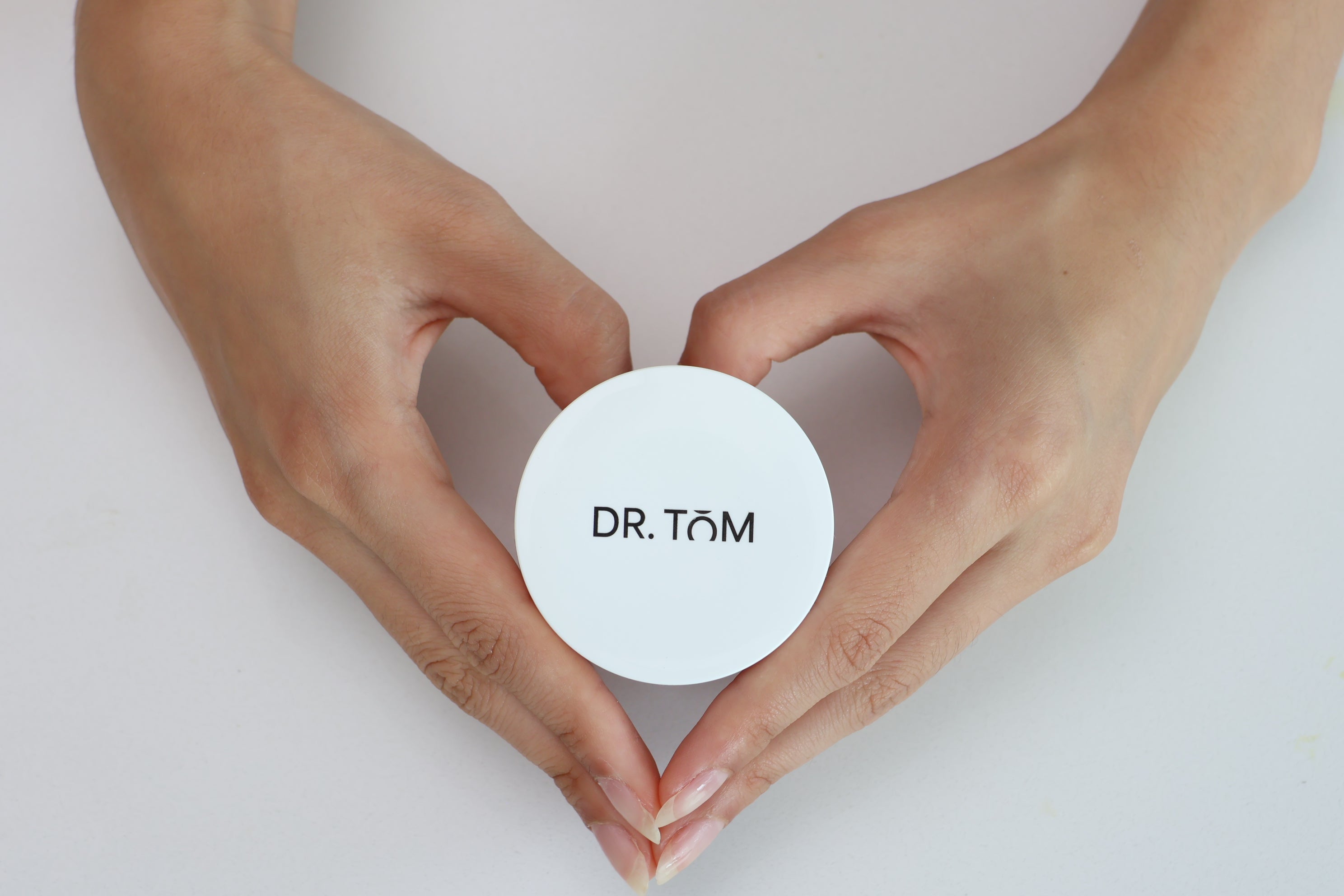 A top-down view of a person's hands holding a round white cosmetic jar with the brand name 'DR. TÖM' printed in black on the lid, set against a clean, light gray background.